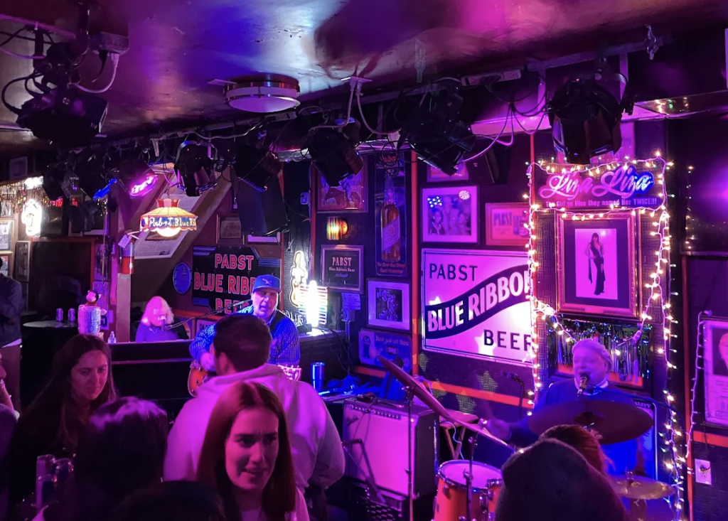 Live band performing inside Bob & Barbara's Lounge, lit in purple with string lights and Pabst Blue Ribbon memorabilia on the walls.
