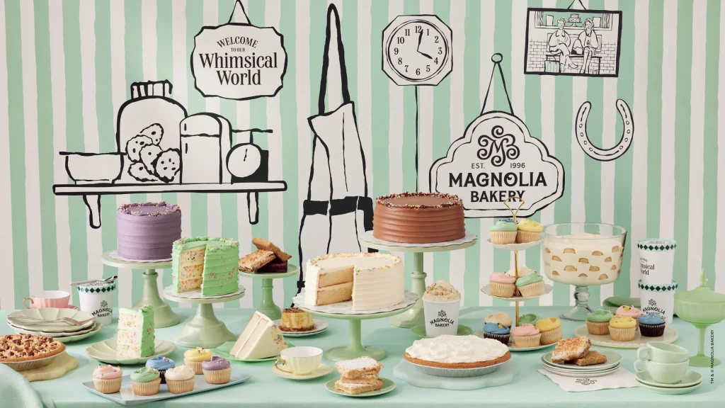 Magnolia Bakery display table loaded with layer cakes, cupcakes, cheesecakes and banana pudding, set against a mint-striped backdrop with illustrated bakery shelves and the Magnolia Bakery logo