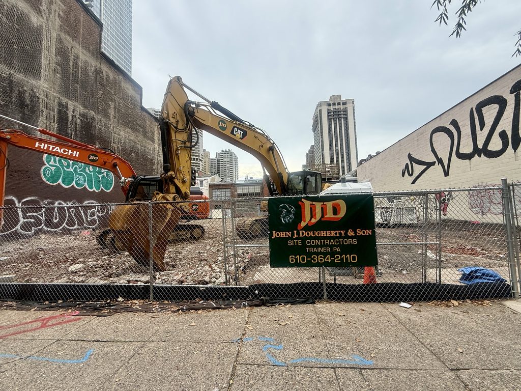 Yellow Caterpillar and orange Hitachi excavators on the rubble-covered lot at 2012 Chestnut Street, behind a chain-link fence with a John J. Dougherty & Son Site Contractors sign.