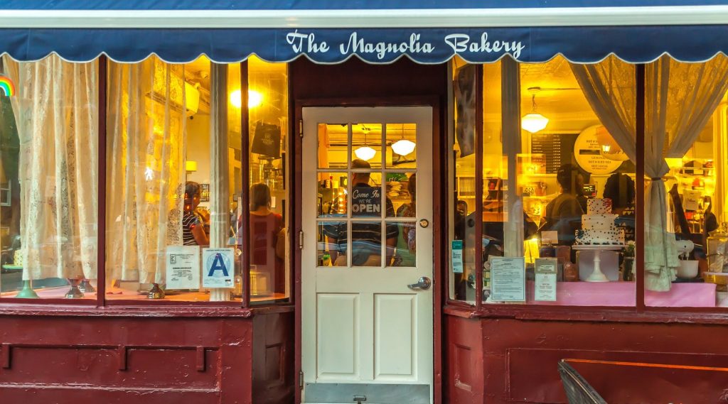 The original Magnolia Bakery storefront in New York’s West Village, with a blue scalloped awning and warm light glowing through the windows