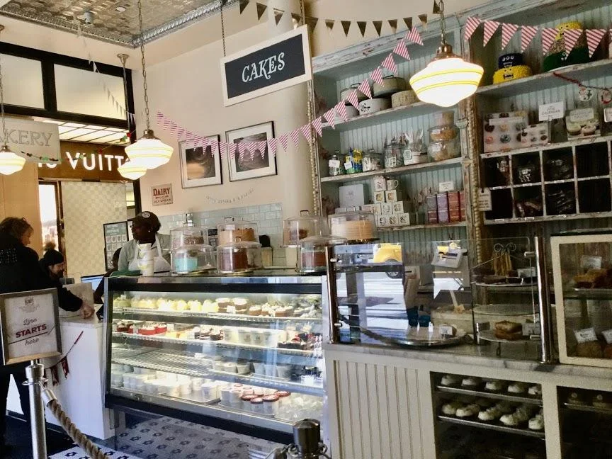 Inside a Magnolia Bakery, with a glass case full of cupcakes and cakes, pink and green bunting, a CAKES sign, and shelves of baked goods